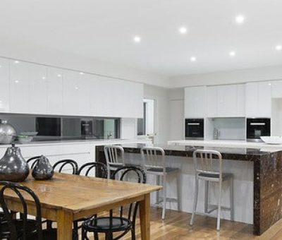 A modern kitchen with white cabinets, a marble island with bar stools, and a wooden dining table with black chairs, all under bright ceiling lights.