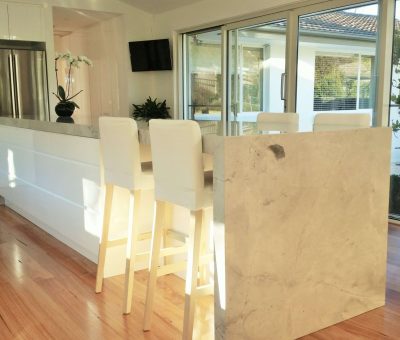 Bright kitchen with a large marble island, three white cushioned stools, hardwood floors, and sliding glass doors offering a view of greenery outside.