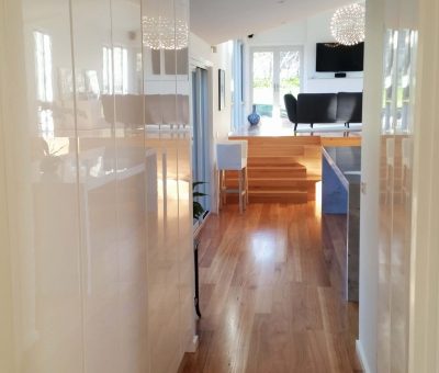 Bright hallway with wooden floors leading to a sunlit living area. Modern decor includes a large chandelier, black sofa, and a wall-mounted TV.