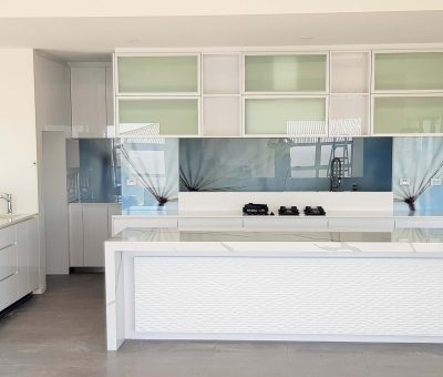 Modern kitchen with sleek, white cabinetry and a large central island. Glass backsplash with subtle blue tones adds elegance. Minimalist and bright ambiance.