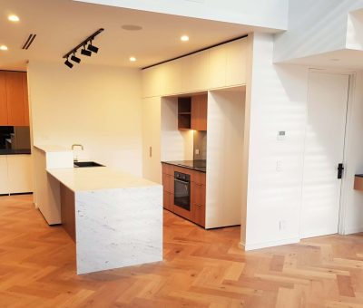 Modern kitchen with light wood herringbone flooring, marble island, and sleek cabinetry. Spotlights and large windows create a warm, inviting atmosphere.