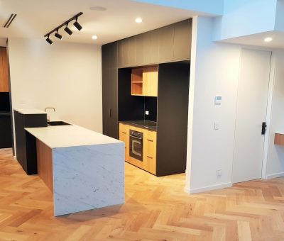 Modern kitchen with herringbone wooden floors, marble island, black cabinets, and built-in oven. It features track lighting and wooden accents.