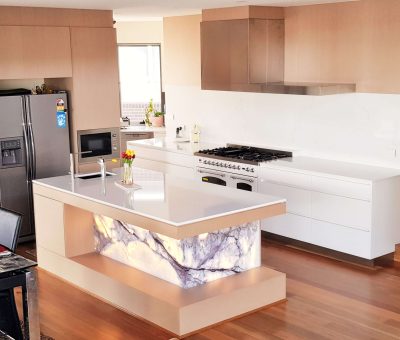 Modern kitchen with a sleek design features a white island with a marbled illuminated panel, stainless steel appliances, and light wood cabinetry.