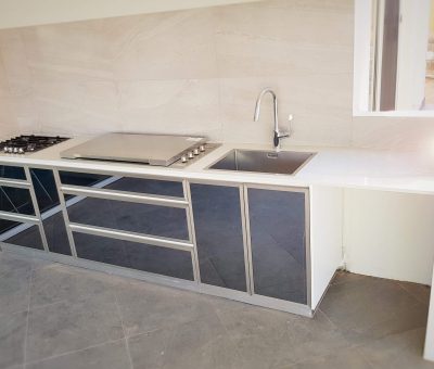 Modern outdoor kitchen with sleek gray cabinets, a gas stove, and a large sink under a window. Bright and minimal design, creating a clean ambiance.