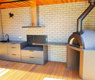 Modern outdoor kitchen with a sleek grill, sink, and brick oven. The area features wood flooring, brick walls, and a wooden ceiling, creating a warm atmosphere.