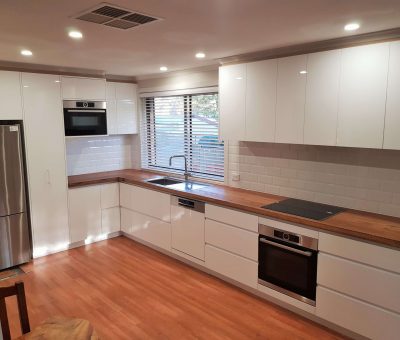 Modern kitchen with white cabinets, wood countertops, and stainless steel appliances. Features a sink under a window with blinds, and warm lighting.