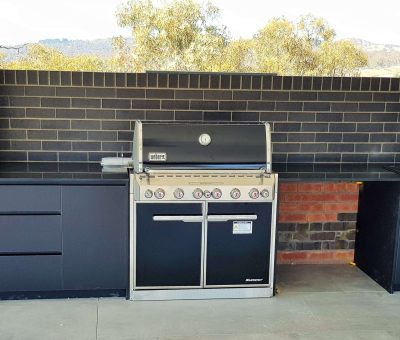 Modern outdoor kitchen with a sleek black grilling station, adjacent black countertops, and built-in sink. Brick wall backdrop and trees visible outside.
