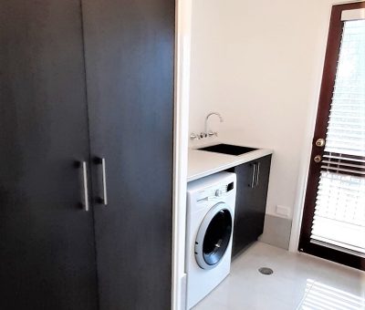 Laundry room with glossy white tile floor, dark cabinets, and a front-loading washer beneath a black countertop. A door with a window is on the right.