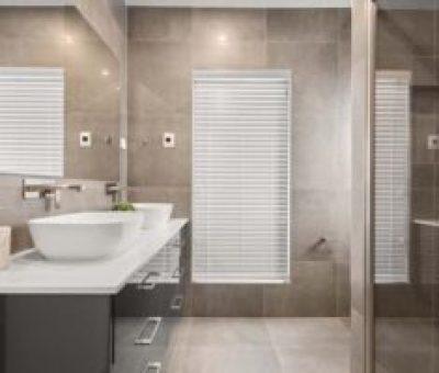 Modern bathroom with a white sink, faucet, and large mirror. Beige tiles and closed blinds on window create a clean, serene ambiance.