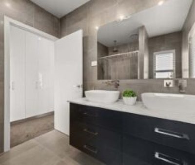 Modern bathroom with a sleek gray design. Features double sinks, a large mirror, and a small plant. Shower area visible through an open door.