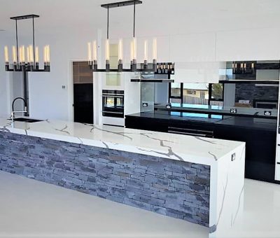 Modern kitchen with sleek design, featuring a long marble island with stone accents, dual chandeliers, glossy white and black cabinets, and a shiny floor.