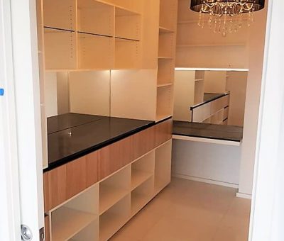 Modern walk-in closet with white and wood shelving, black countertops, and a mirrored wall. A black chandelier adds elegance to the bright space.