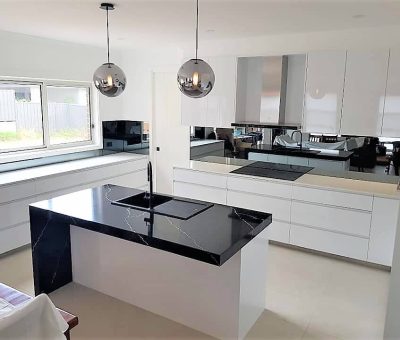 Modern kitchen with white cabinetry and black marble countertops. Central island features a sink. Two metallic pendant lights hang above, and a window allows natural light. Sleek, minimalist design.