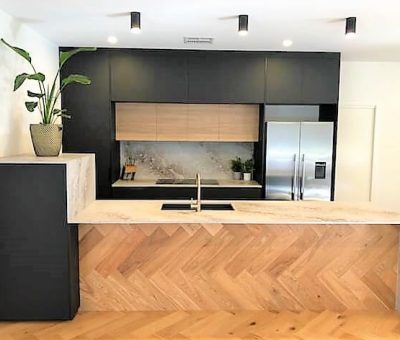 Modern kitchen with a sleek, minimalist design. Features a light wood herringbone island, black cabinets, stainless steel fridge, and a potted plant.
