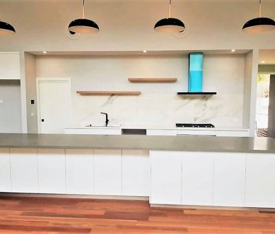Modern kitchen with white cabinets, gray countertop, and wooden floor. Pendant lights hang above, and a blue range hood adds a pop of color. Minimalist design.