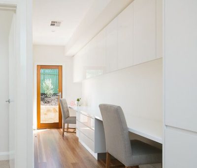 Bright, minimalist home office with white cabinets and desk, gray chair, and a wooden floor. A door with a large window lets in natural light, creating an airy feel.