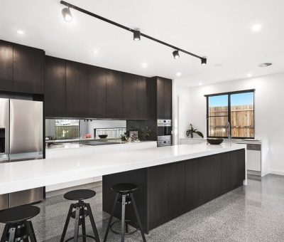 Sleek modern kitchen with dark cabinets, long white island, and stainless steel appliances. Three black stools, bright lighting, and a large window.