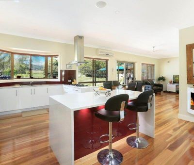 A spacious, modern kitchen with a central island and bar stools is connected to a cozy living area. Large windows offer a view of green scenery, and a warm, inviting fireplace adds charm.