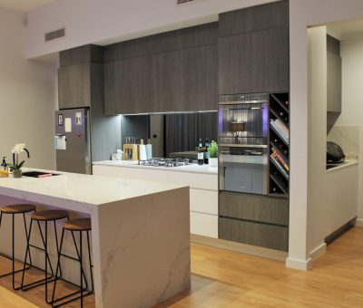 Modern kitchen with dark cabinets, marble island, and wooden floor. Features include an oven, wine rack, and three stools. Minimalist and sleek design.