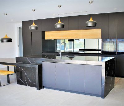 Sleek modern kitchen with black cabinetry, marble countertops, and light wood accents. Pendant lights hang above a central island and bench seating.