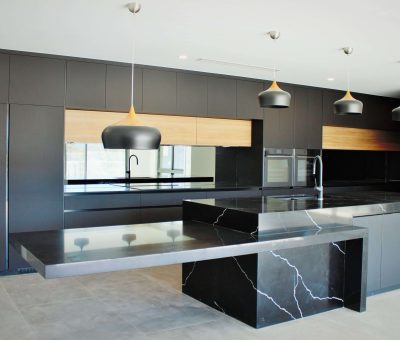 Modern kitchen with sleek black cabinets, a marble island, pendant lights, and wooden accents. The space exudes a luxurious and minimalist design.