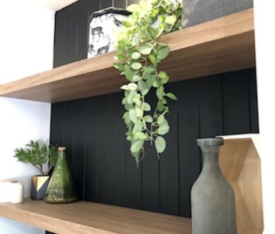 Two wooden shelves against a black panel wall hold decor items: a potted plant, grey vase, and storage box. A hanging vine adds a touch of nature.
