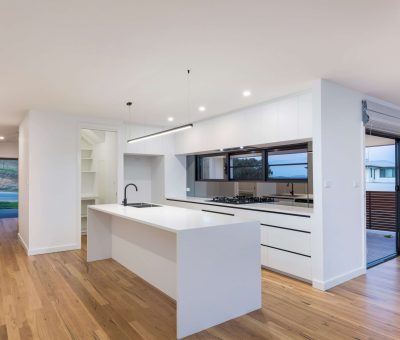 Modern kitchen interior with white island, sleek cabinetry, and wood flooring. Natural light enters through large windows and a sliding glass door.