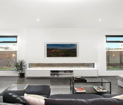 Modern living room with a sleek design, featuring black couches, a glass coffee table, and minimal decor. Large windows and a wall-mounted TV create an open, airy feel.