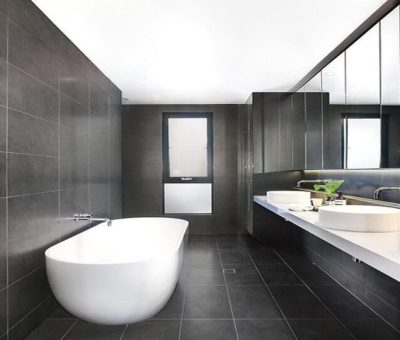 Modern bathroom with dark tiled walls and floors, white freestanding bathtub, and large mirrored vanity.