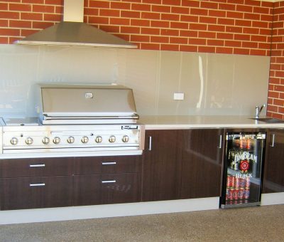Outdoor kitchen with a stainless steel grill, dark wood cabinets, a mini fridge stocked with drinks, placed against a red brick wall. Modern and inviting.