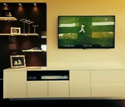 Modern living room with a wall-mounted TV showing a sports game. Below is a beige cabinet with shelves, photos, and decor items, lit by spotlights. Cozy and stylish ambiance.