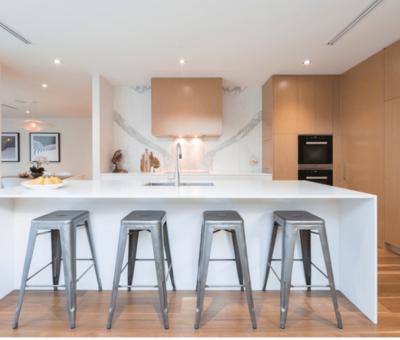 Modern kitchen with a large white island, four gray metal stools, wooden cabinets, and a marble backsplash. Bright and minimalist atmosphere.