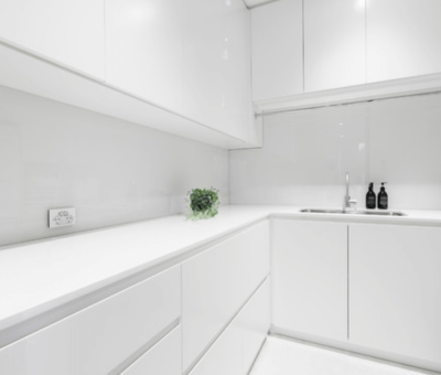 A sleek, minimalist kitchen with glossy white cabinets and countertops. A small potted plant and two black soap dispensers add contrast to the clean design.