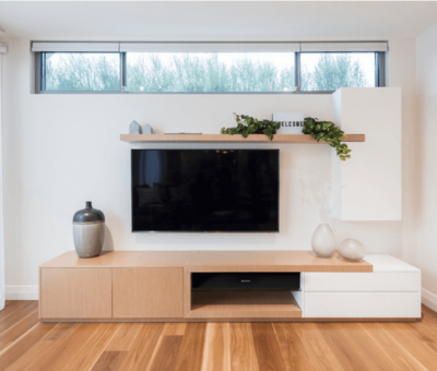 Modern living room with a sleek wooden TV stand, decorative vase, and greenery on shelves. A large flat-screen TV is centered on the wall. Bright and minimalistic ambiance.