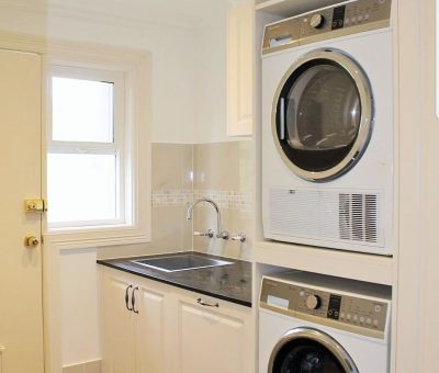 A compact, modern laundry room with a stacked washer and dryer, beige cabinetry, stainless steel sink, and a window providing natural light.