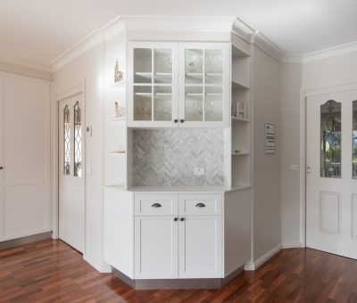 A bright kitchen corner with white cabinetry, glass-paneled upper cupboards, and a herringbone-tiled backsplash. Hardwood floors add warmth to the space.