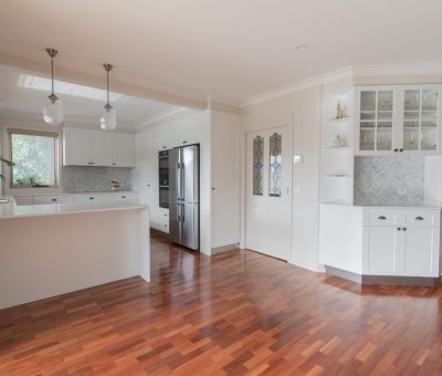 A modern kitchen with wood flooring, white cabinets, and a sleek stainless steel fridge. Pendant lights hang over the countertop, creating a bright, airy feel.