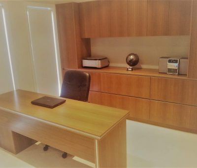 Modern office with wooden desk and black chair. A laptop sits on the desk, with a printer, globe, and stereo on wooden shelves behind, creating a tidy, professional atmosphere.